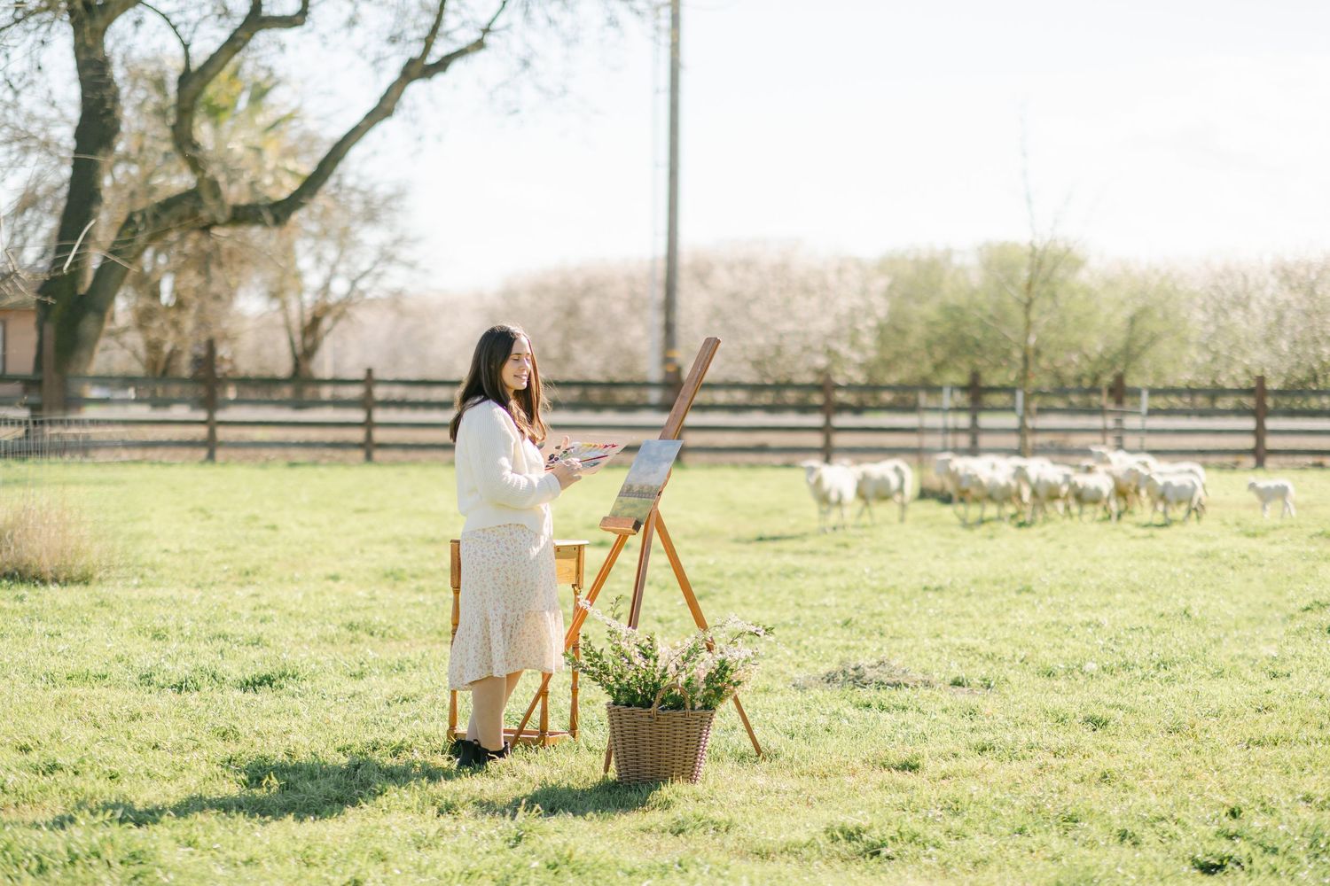 A pregnant woman painting on an easel in a sunny farm field with grazing sheep and wooden fence in background.