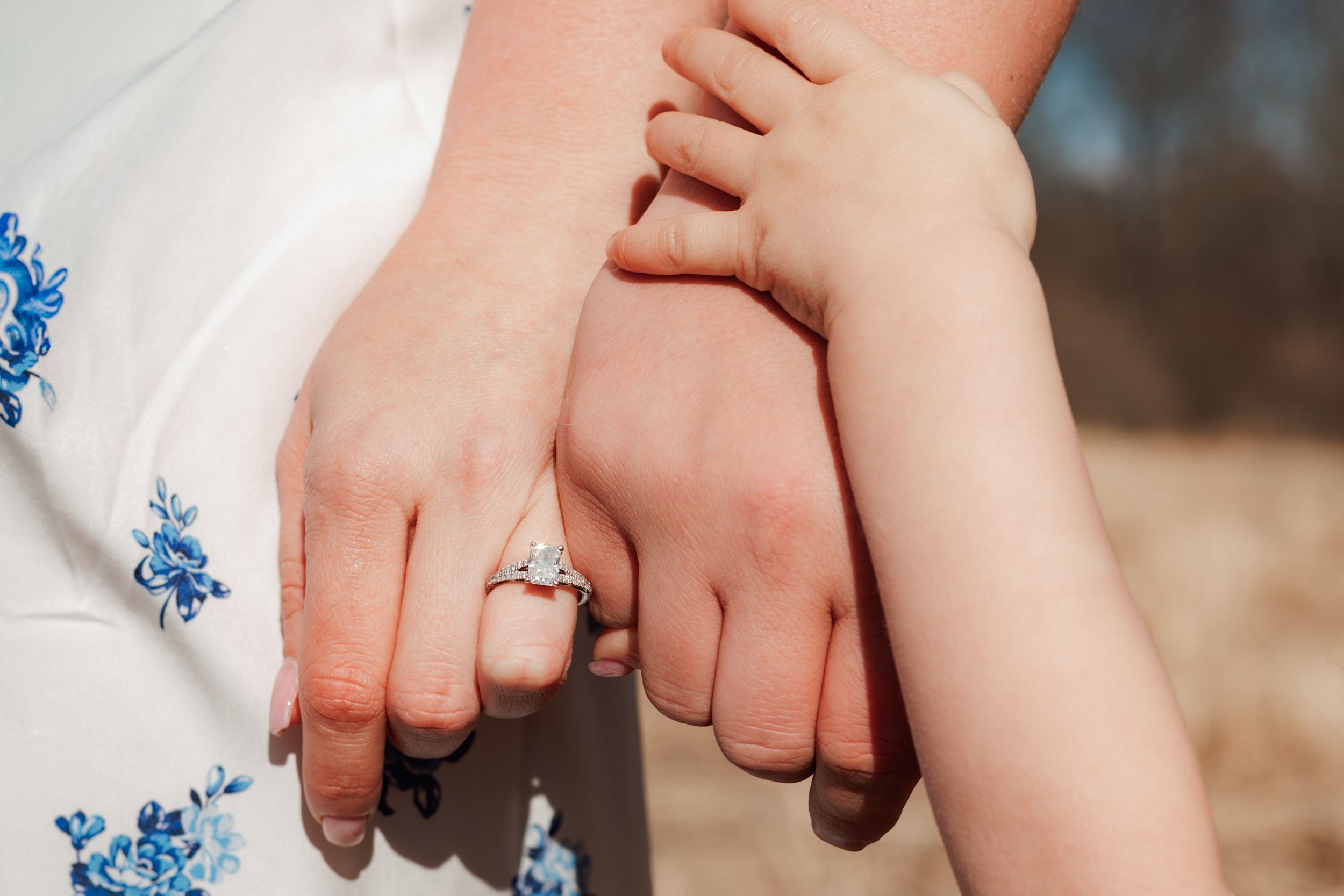 Close-up of a woman's hand wearing a diamond engagement ring, with a child's hand resting on her arm.