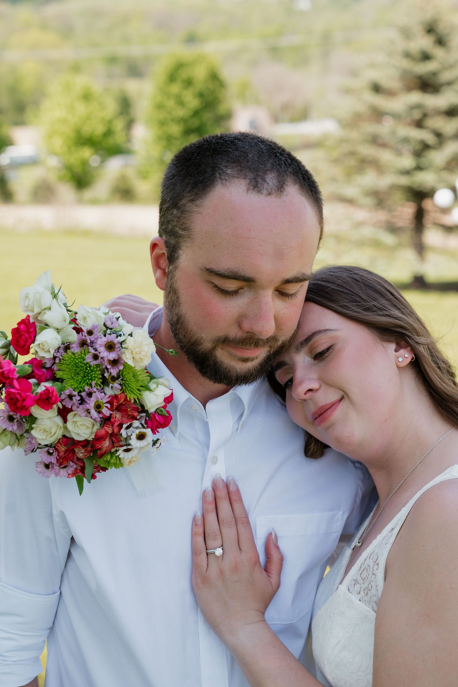 Newlywed couple embracing outdoors, bride resting head on groom's shoulder holding colorful wedding bouquet with eyes closed.