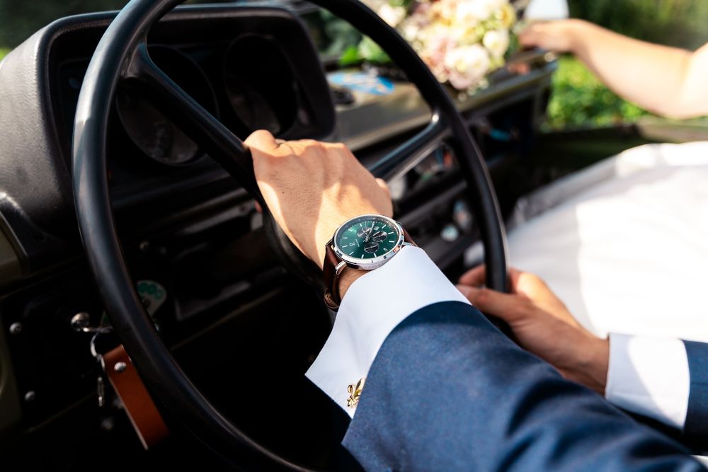 Man in blue suit with green watch driving a vintage car, close-up of hands on steering wheel.