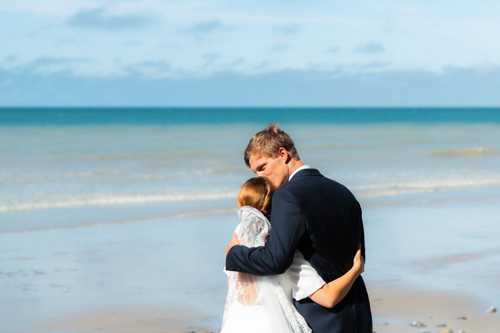 Bride and groom share a romantic embrace on a sandy beach with turquoise ocean waves in the background.