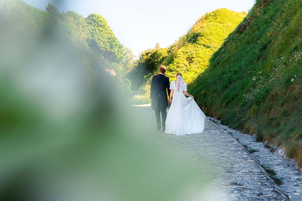 Bride and groom walking hand in hand along a scenic stone path surrounded by lush green hills on their wedding day.