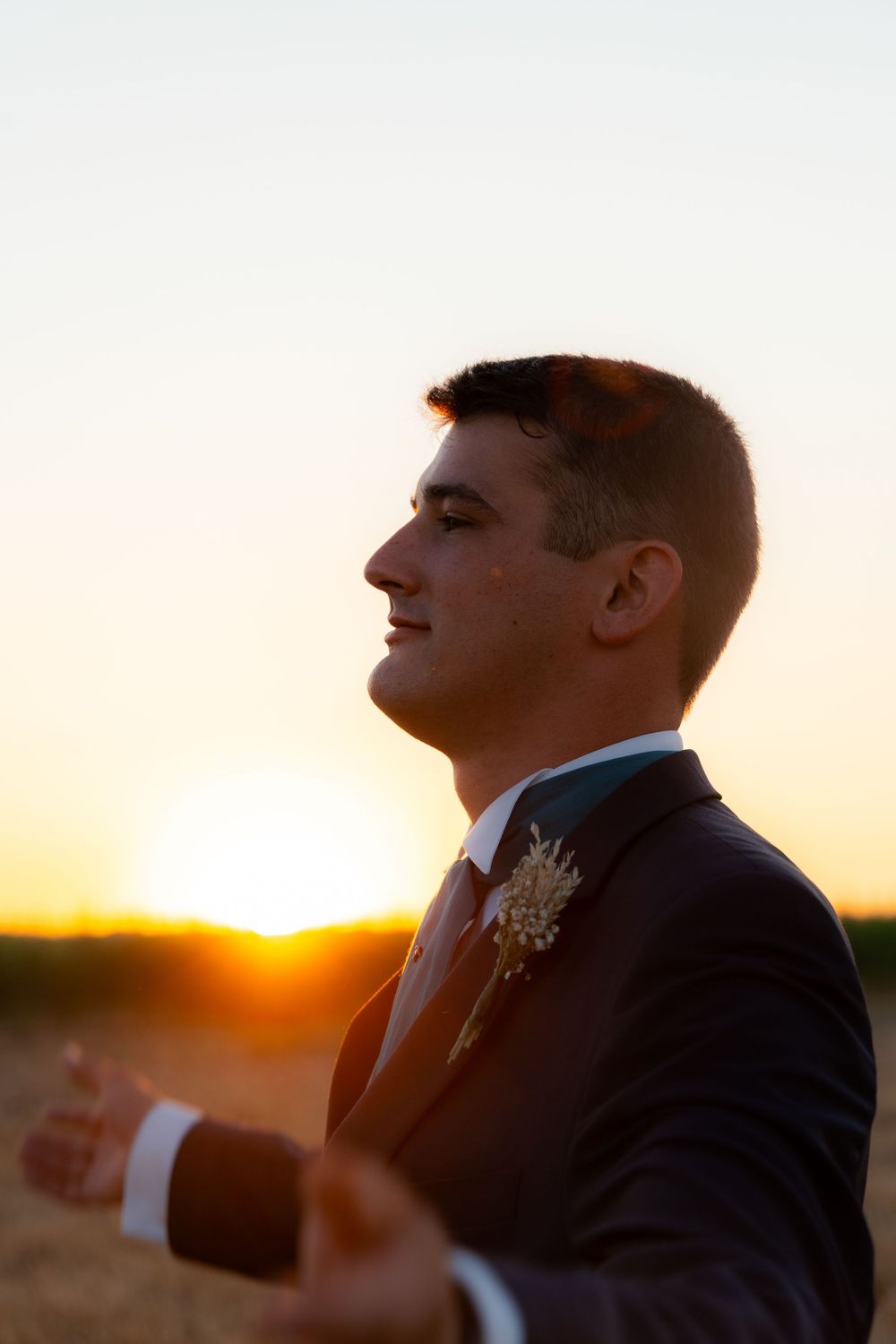 Groom in dark suit with boutonniere standing in profile against golden sunset in an open field.