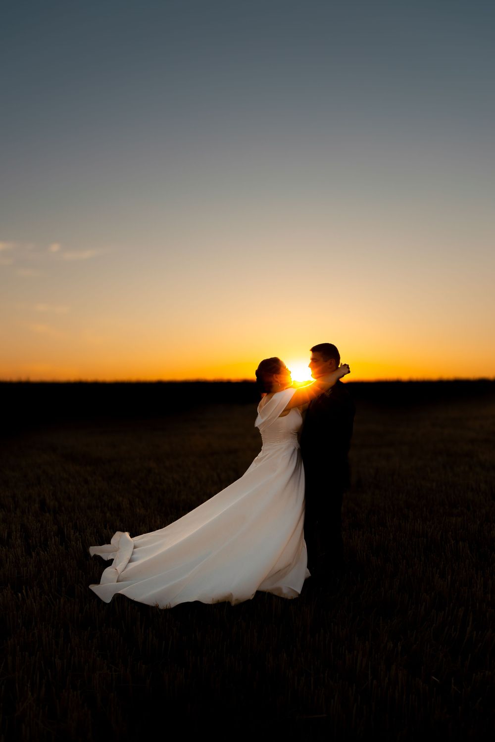 Bride and groom silhouetted against a golden sunset in an open field, wedding dress flowing in the breeze.