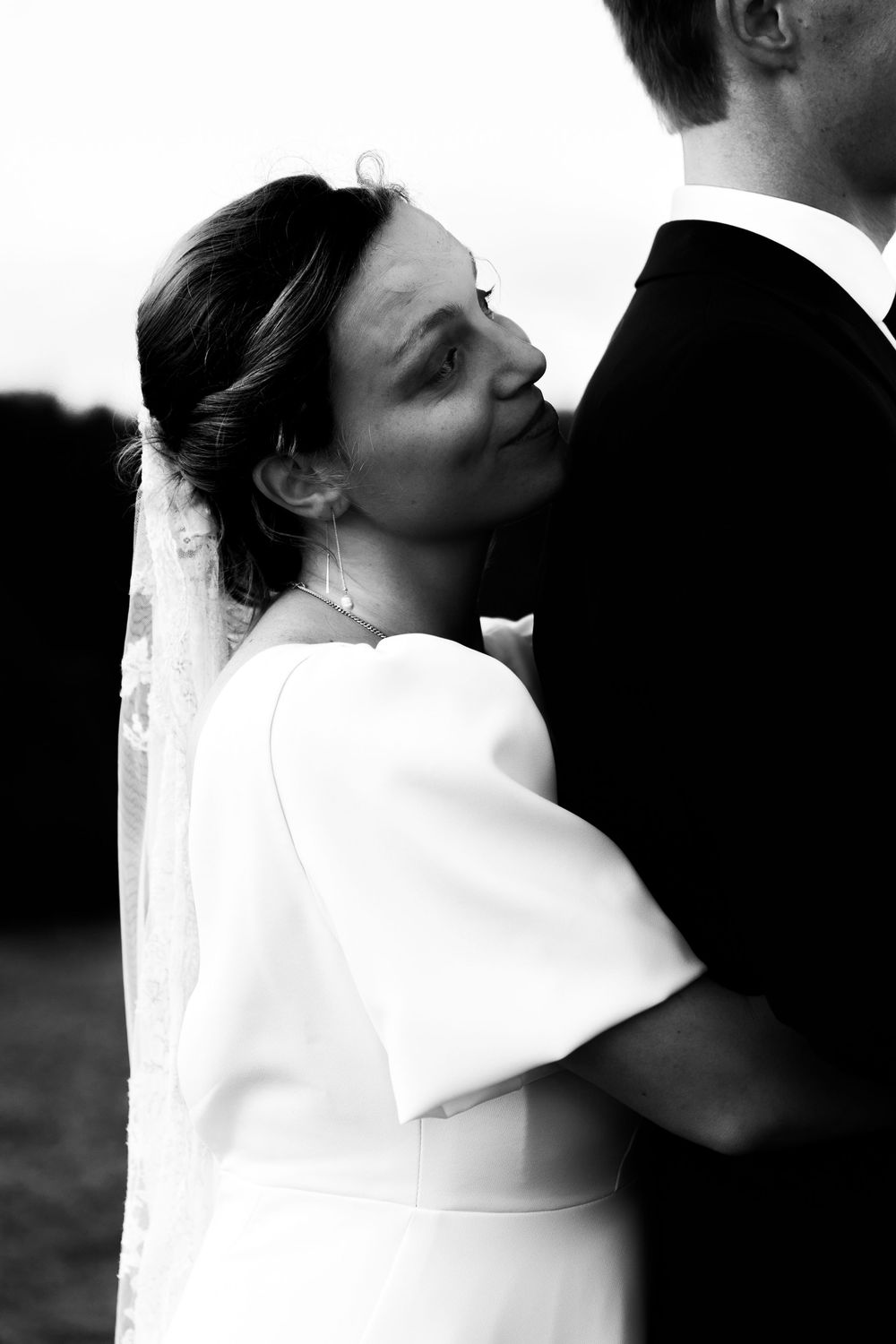 Black and white wedding photo of bride in white dress with veil embracing groom in suit outdoors.