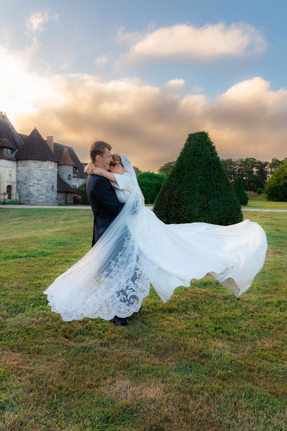 Bride and groom dancing on green lawn before a castle at sunset, white wedding dress flowing in the breeze.
