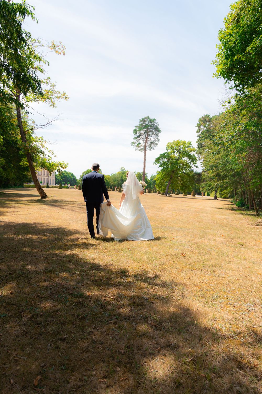 Bride and groom walking hand in hand through a sunny park, bride in white gown with veil trailing on dry grass.