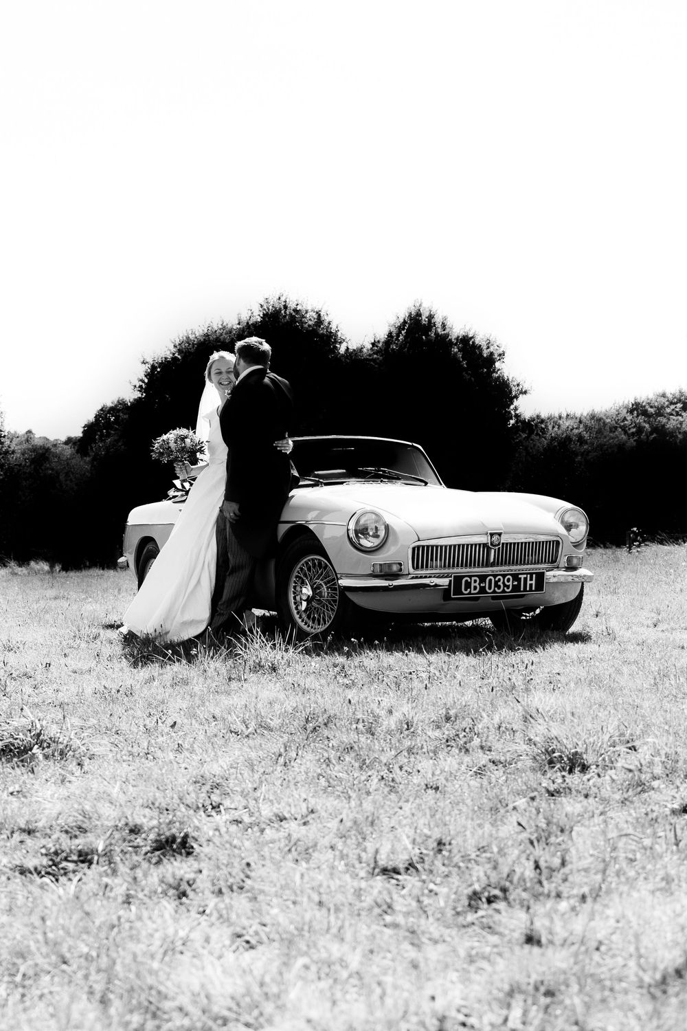 Black and white photo of a bride and groom kissing beside a vintage MG convertible car in a grassy field.