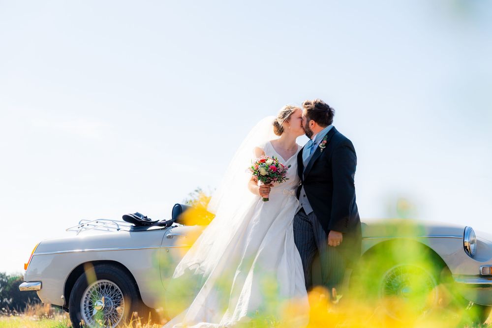 Bride and groom kissing beside a vintage silver convertible car on their wedding day outdoors.