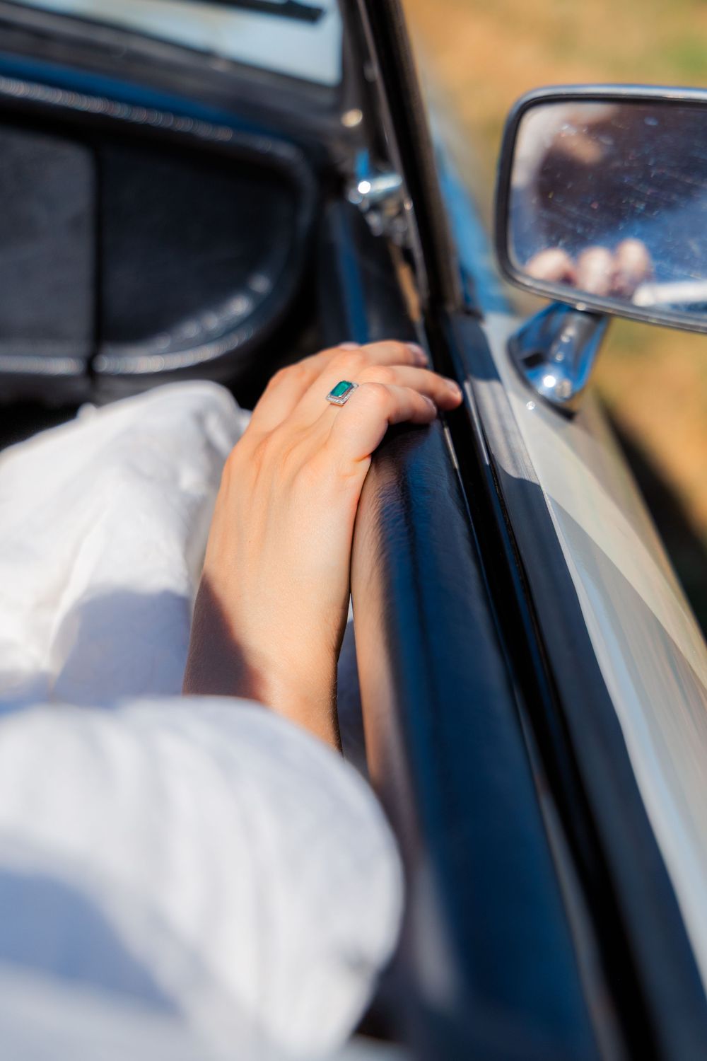 Close-up of a hand with a green gemstone ring resting on a vintage car door, with a side mirror reflecting sunlight.