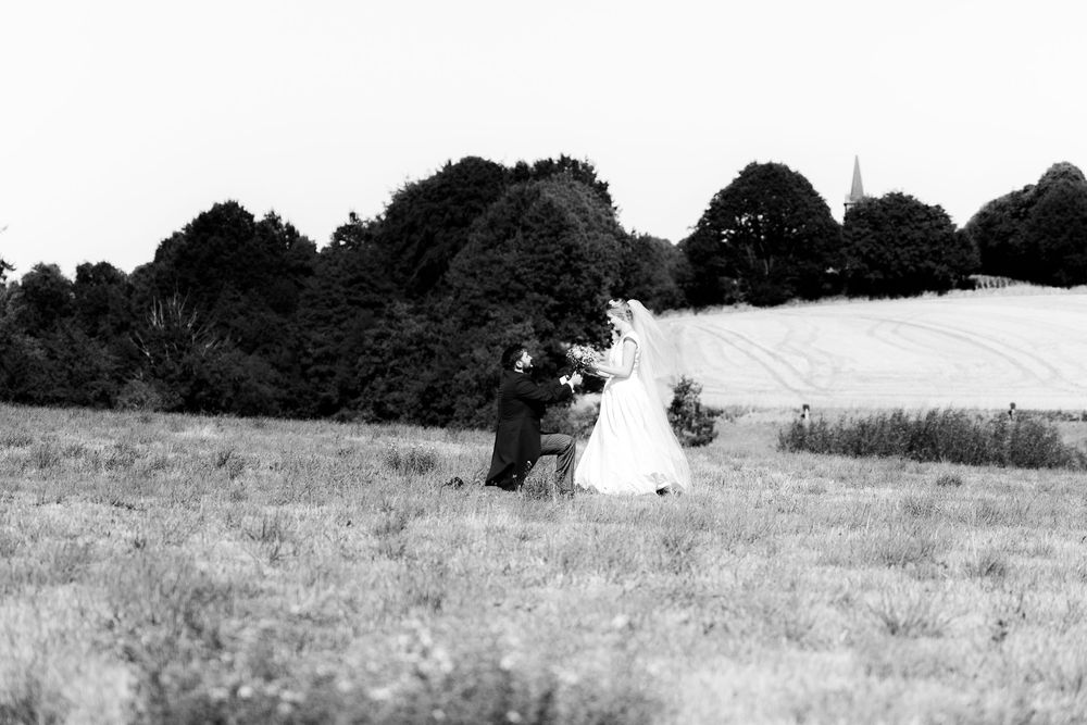 Black and white photo of a bride and groom walking through a field with trees and a church spire in the background.