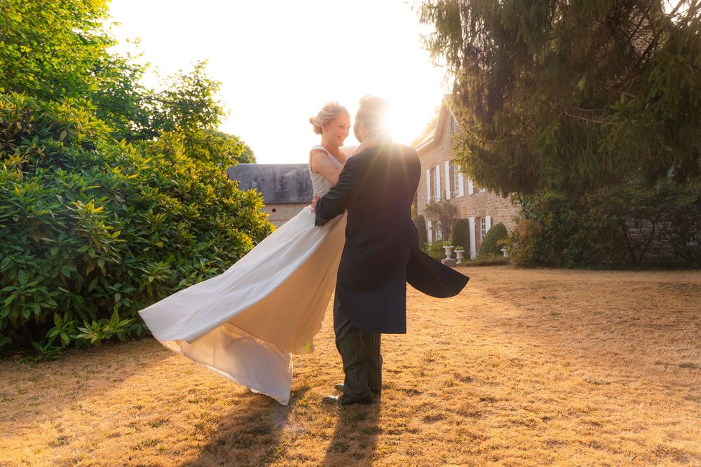Groom lifting bride in flowing white dress at golden sunset outside elegant manor house.