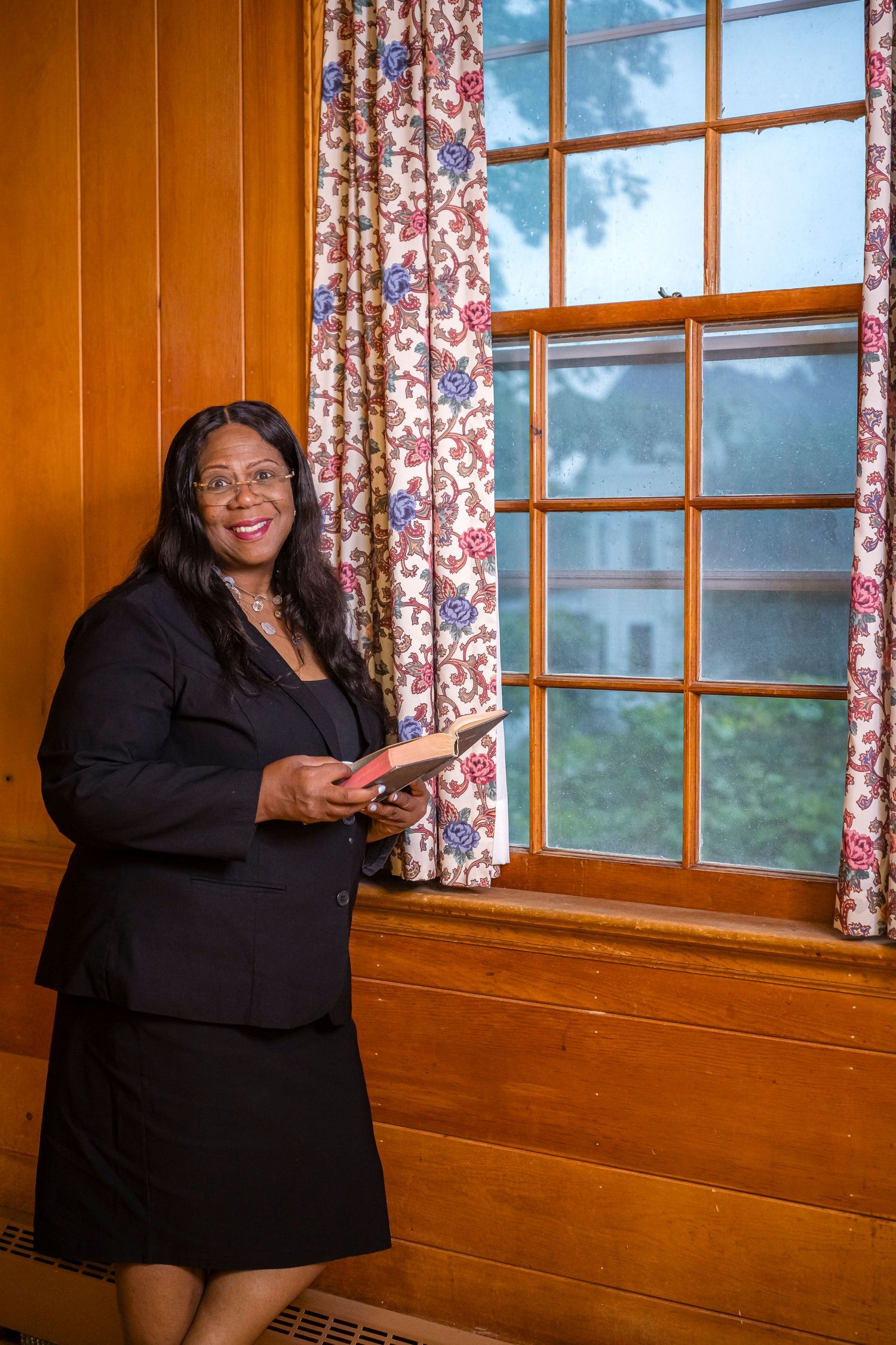 Professional woman in black suit holding a book, smiling beside a wood-paneled wall and floral curtained window.