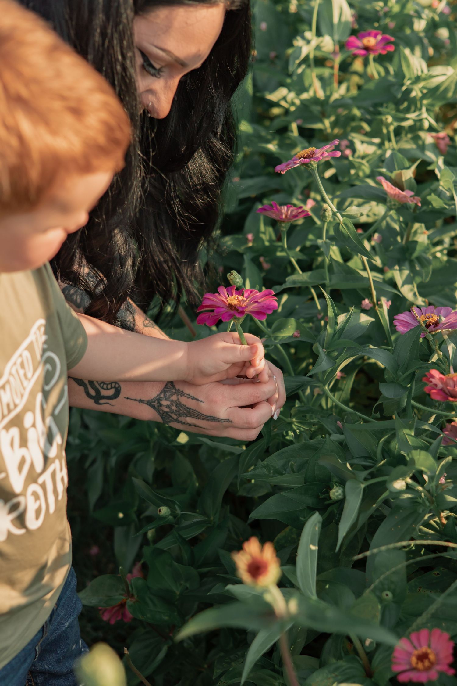 A tattooed woman and red-haired child examine pink zinnia flowers together in a lush garden.