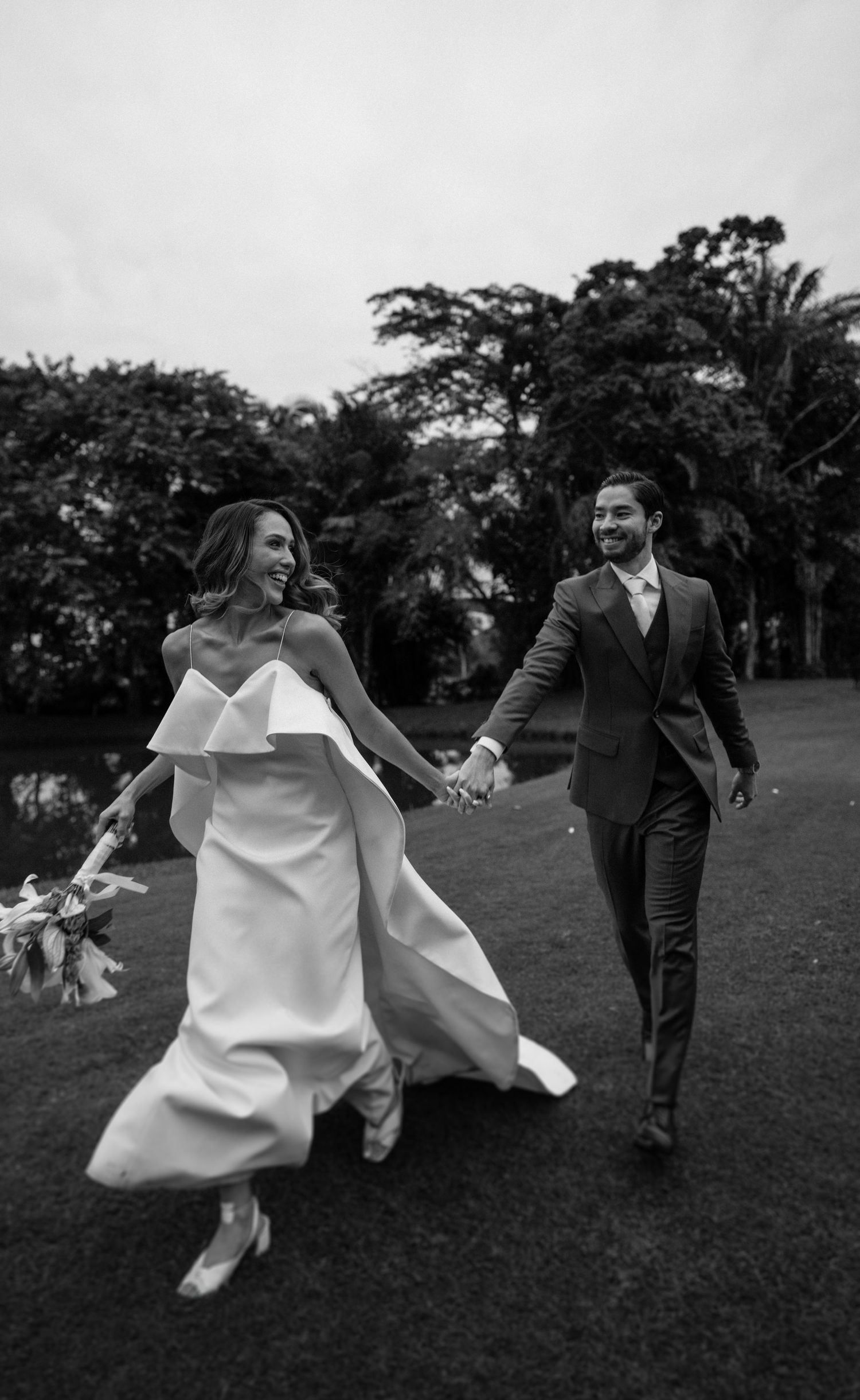 Black and white photo of a smiling bride in a strapless gown and groom in a suit holding hands while walking outdoors.