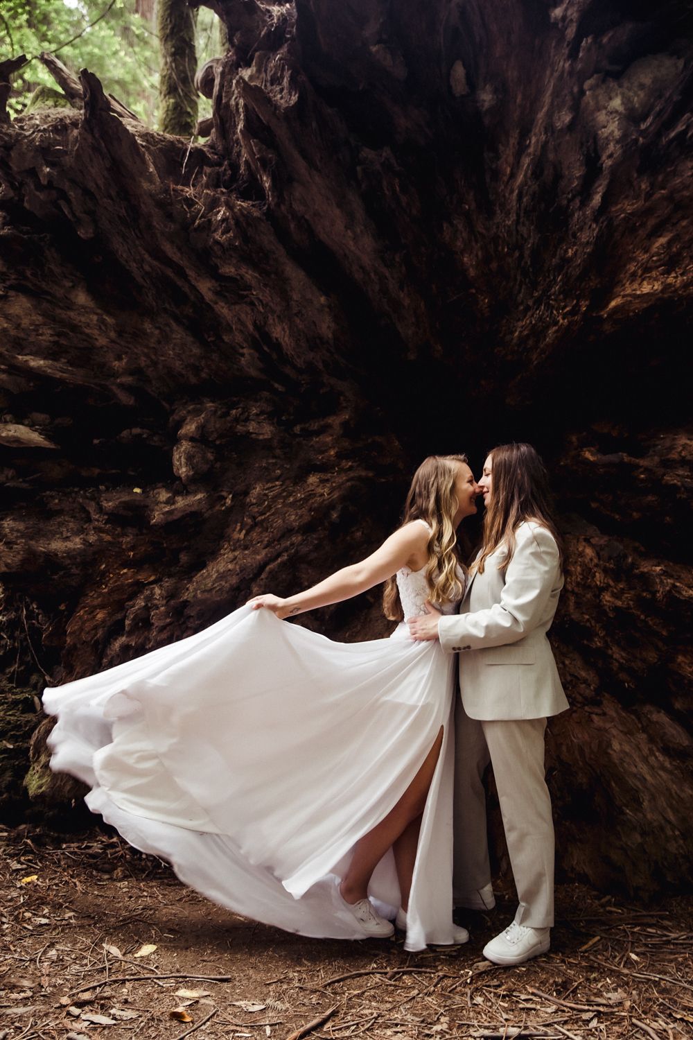 Two brides, one in a flowing white dress, pose in front of a massive redwood tree