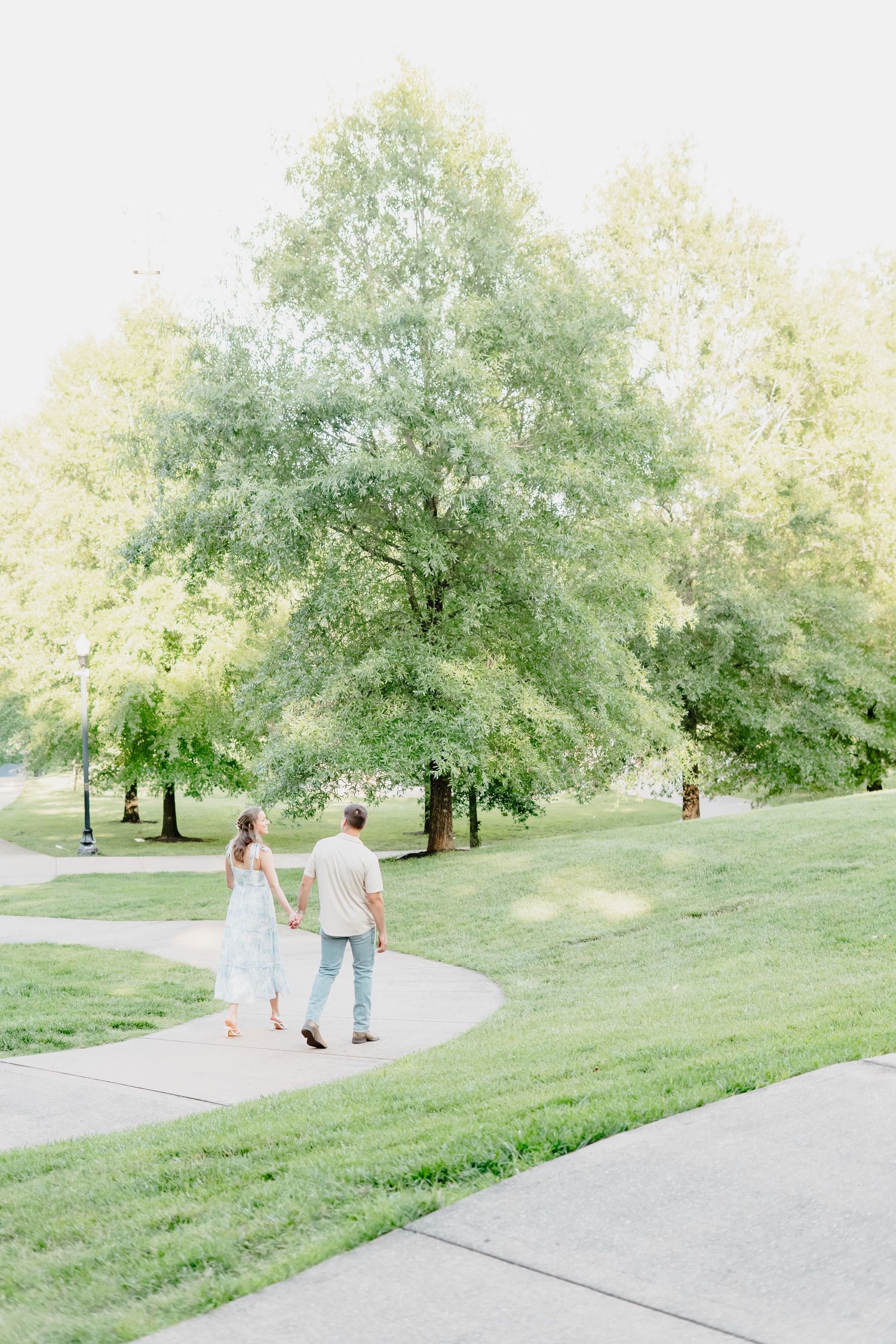 A couple holds hands walking along a curved path in a lush green park surrounded by tall trees.