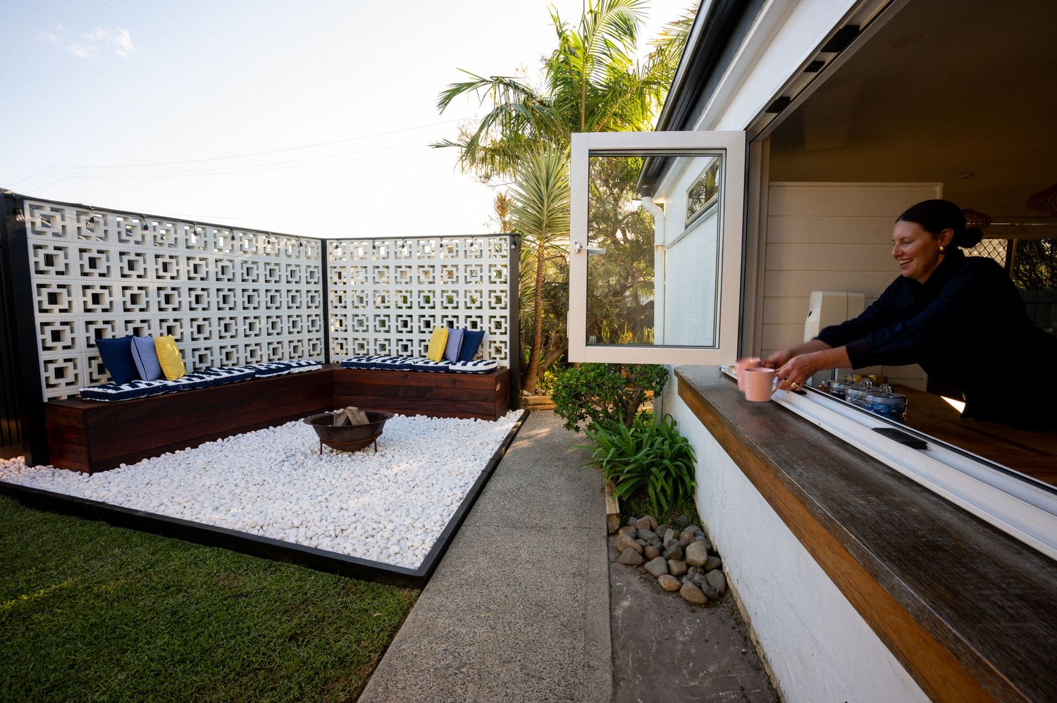 Woman enjoying a modern outdoor patio with white gravel, decorative breeze block wall, and blue cushioned seating area.