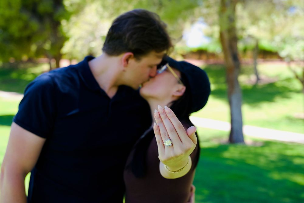 Couple sharing a kiss in a green park, woman displaying an engagement ring on her hand.