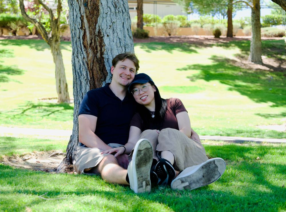 Happy couple sitting together against a tree trunk on green grass in a sunny park.