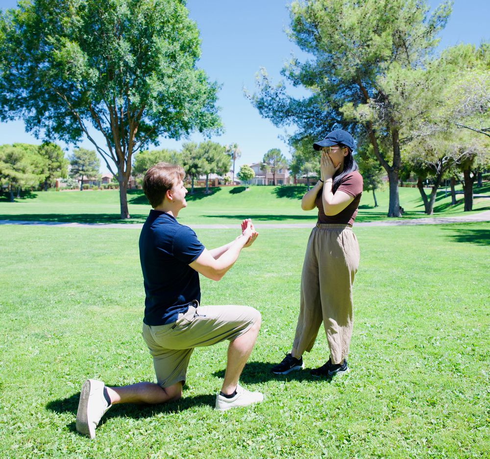 Man down on one knee proposing to his girlfriend in a sunny green park.