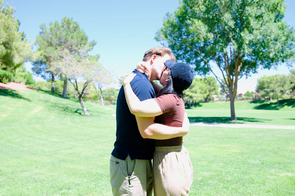 Couple sharing a warm embrace outdoors on a sunny day in a lush green park with trees in the background.