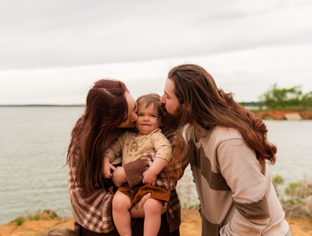 Two parents with long hair kissing their toddler on the cheeks outdoors near a scenic waterfront.