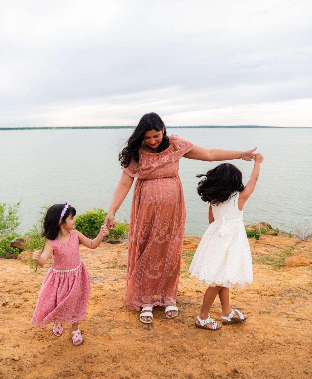 Pregnant woman in pink lace dress dancing with two young daughters on a red clay bluff overlooking a lake.