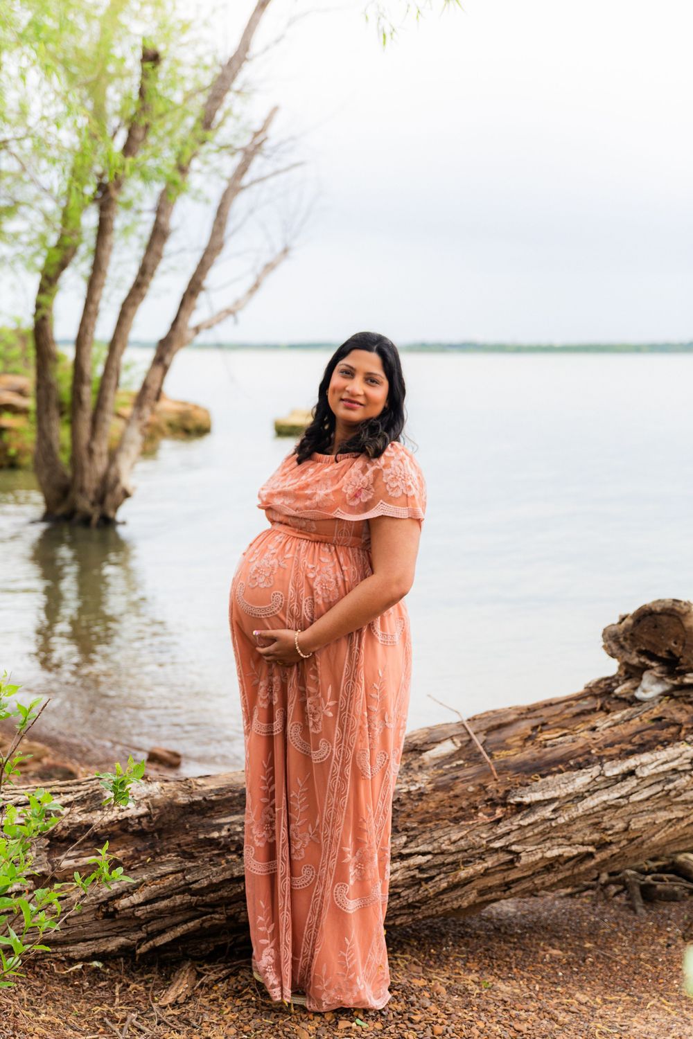 Pregnant woman in pink lace dress cradling baby bump by lakeside with trees and water in background.