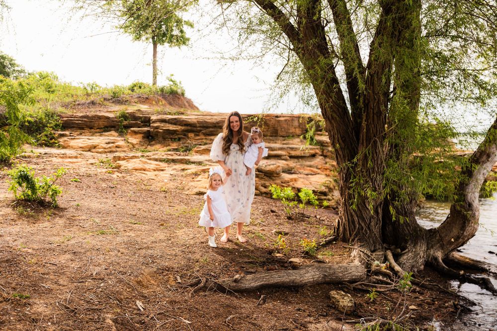 A mother and young daughter in matching white floral dresses stand near a large tree by rocky cliffs outdoors.
