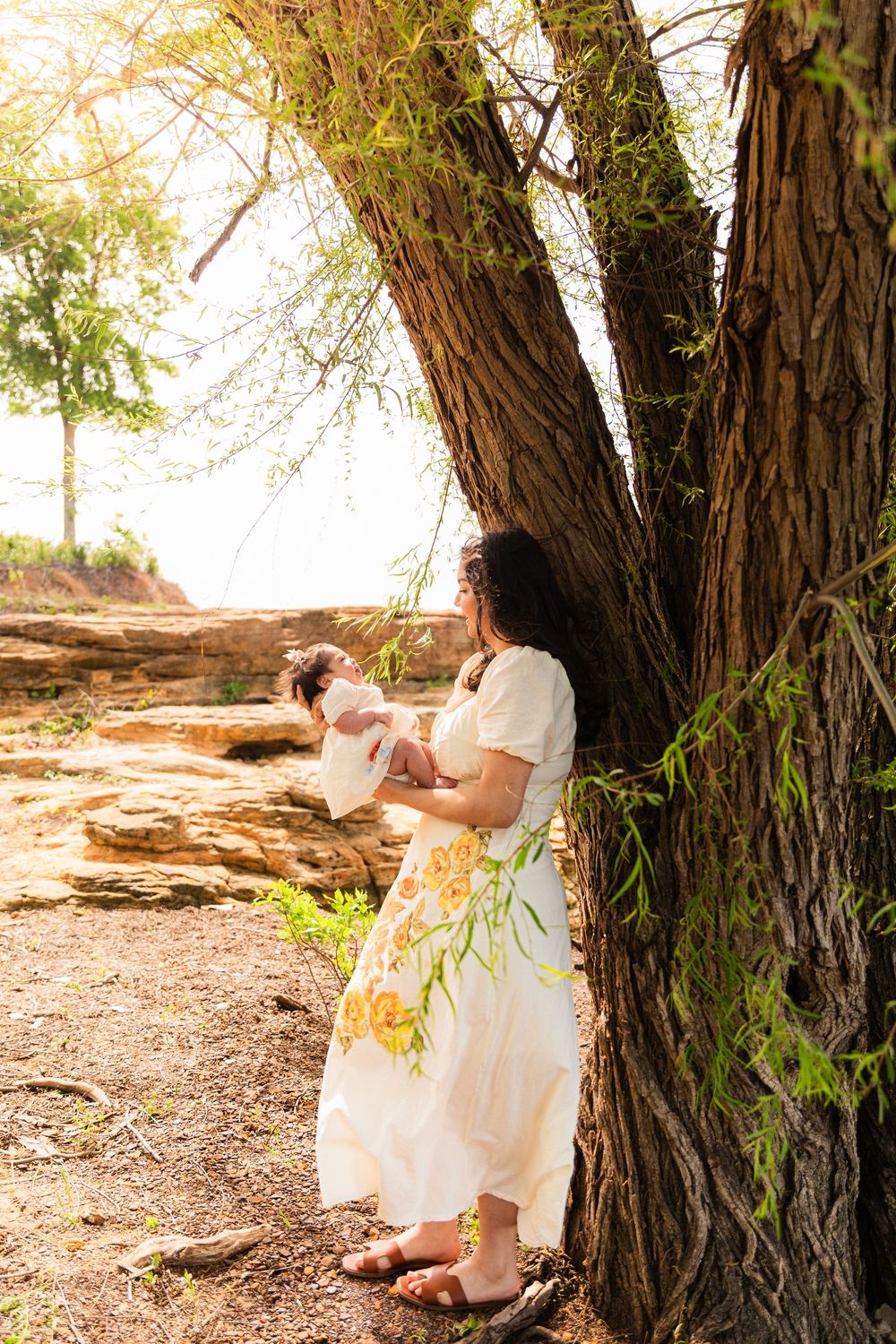 A woman in a floral white dress holds a baby while leaning against a large tree outdoors.