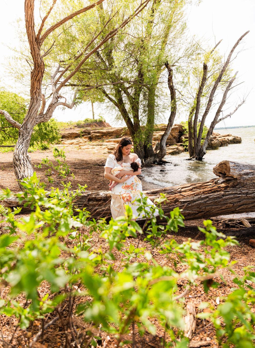 A woman in a floral dress holds a baby near rocky shoreline with willow trees and calm water in a serene outdoor setting.