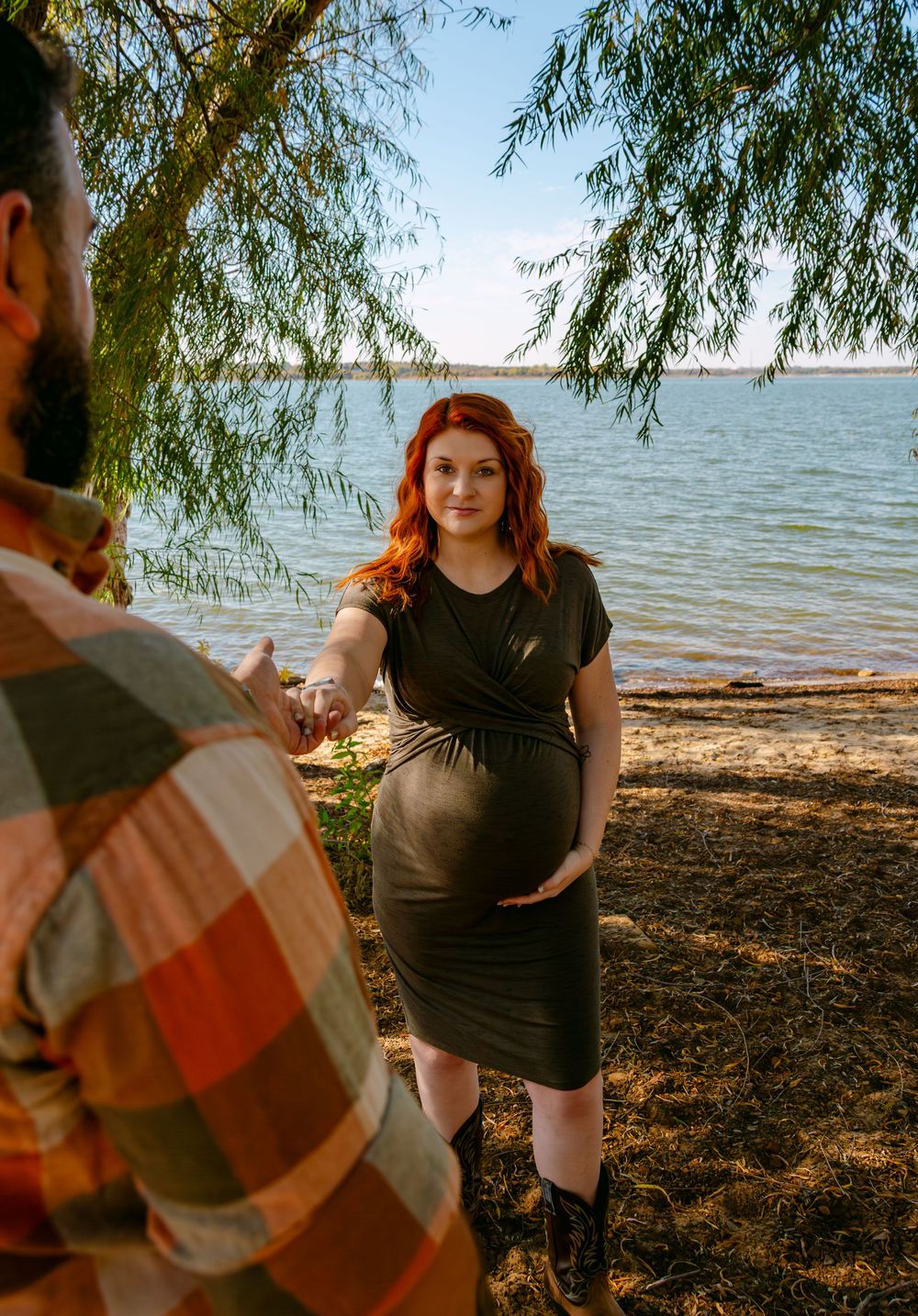 Pregnant redhead woman in olive dress holds hands with bearded man in plaid shirt by a lakeside under willow trees.