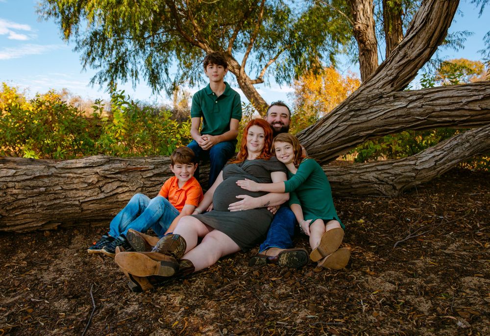 Pregnant woman relaxes against a tree with her family, including three children and a partner, in an outdoor autumn setting.