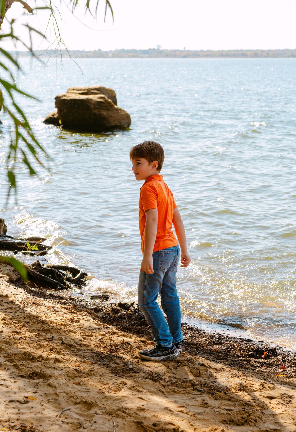 A young boy in an orange shirt and jeans stands on a sandy shore, gazing at calm blue lake waters.