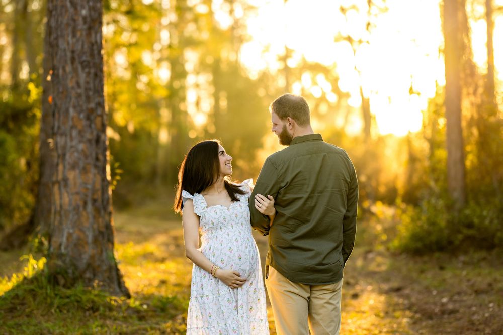 Pregnant couple walking in golden sunlit forest, woman in white floral dress cradling baby bump, man in olive shirt.
