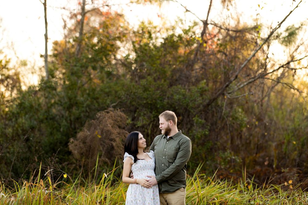 Couple sharing a tender moment outdoors at golden hour, surrounded by lush greenery, in a maternity photo session.