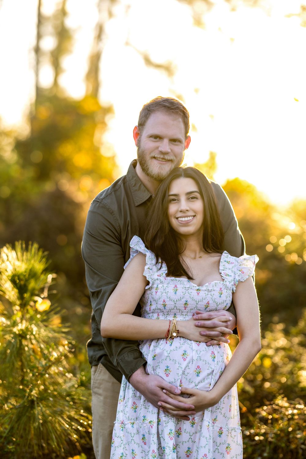 Couple posing outdoors at golden hour, man embracing pregnant woman in floral dress, surrounded by lush greenery.