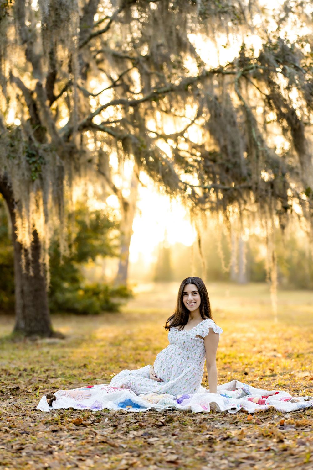 Pregnant woman in white dress sits on colorful blanket under Spanish moss-draped oak trees at golden sunset.