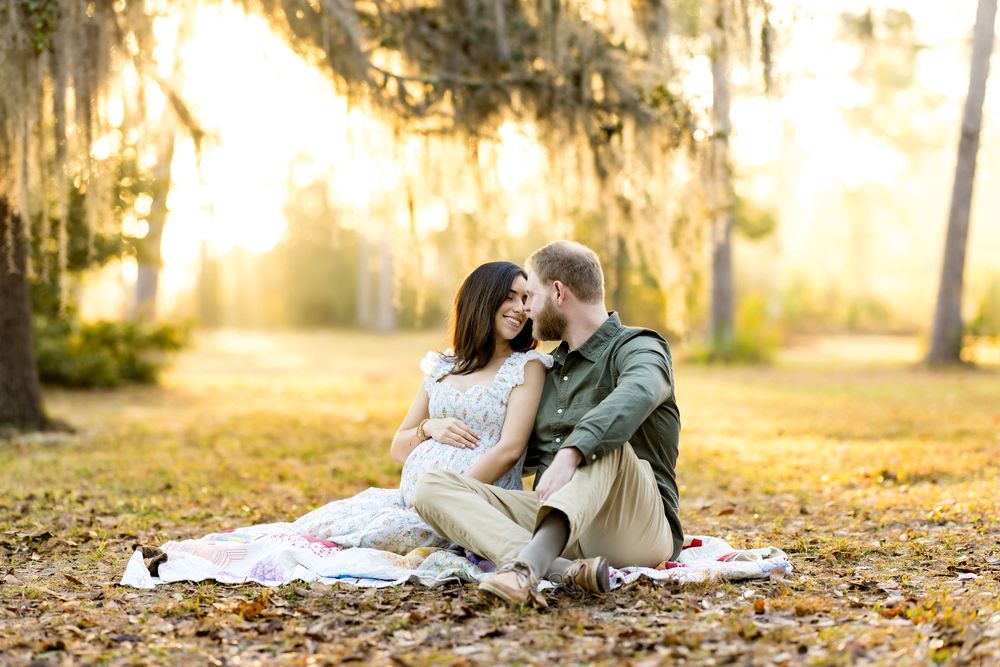 Couple sitting on a blanket in a sunlit park with Spanish moss trees, sharing a tender moment during a maternity photoshoot.
