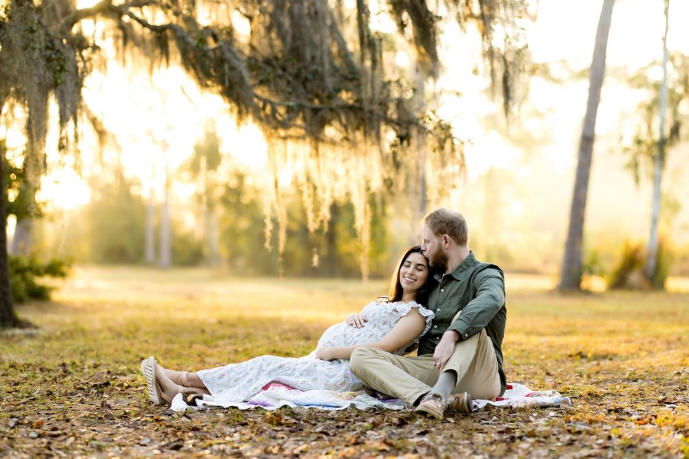 Couple sharing a tender moment on a blanket under Spanish moss trees during a golden hour maternity photo session.