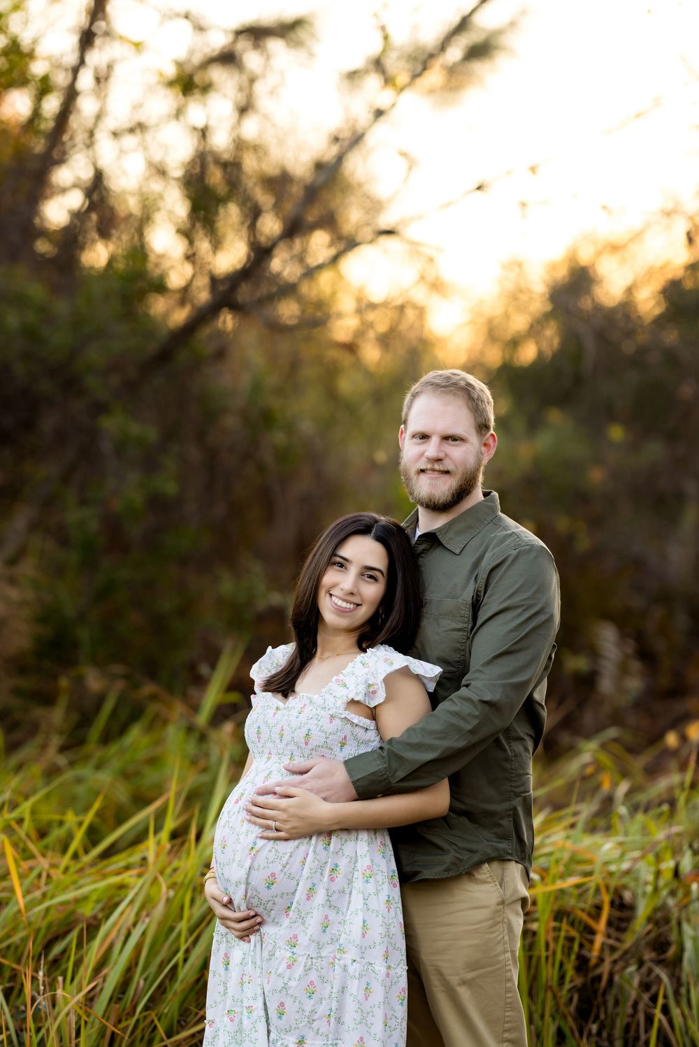 Couple poses outdoors at golden hour, woman in floral maternity dress cradling baby bump, man in green shirt embracing her.
