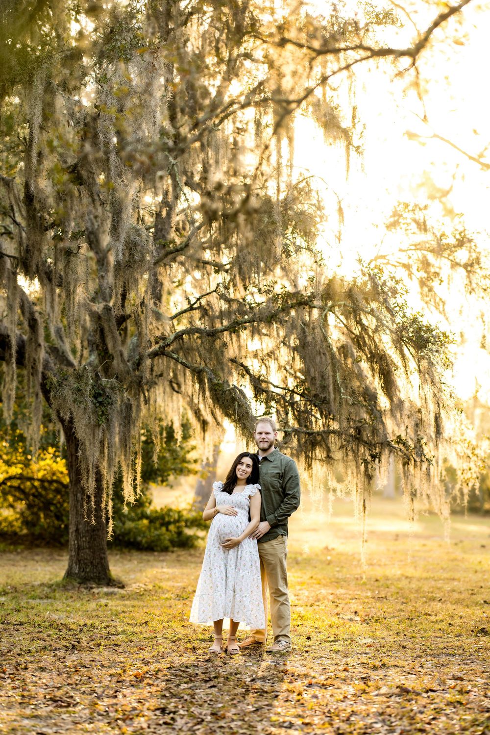 Pregnant couple embracing under Spanish moss-draped oak trees in golden hour light during outdoor maternity photo session.
