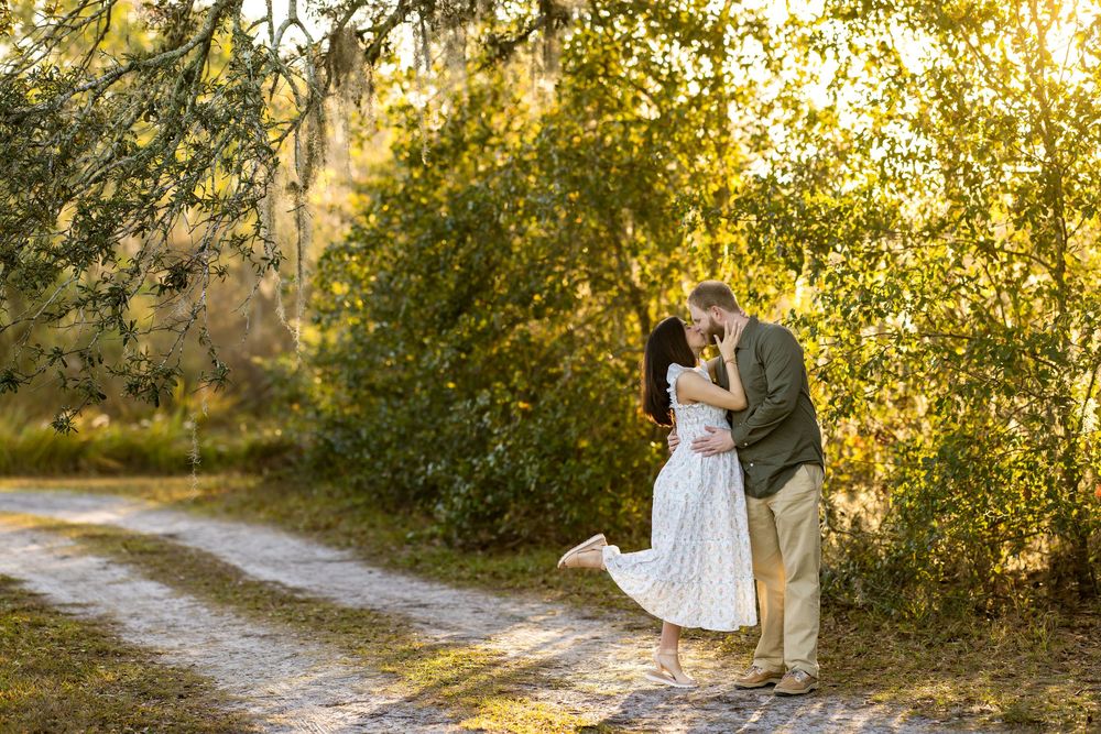 Couple sharing a romantic kiss on a sunlit path surrounded by lush green trees.