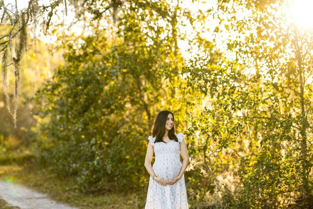 Pregnant woman in white lace dress standing in sunlit park path surrounded by lush green trees.