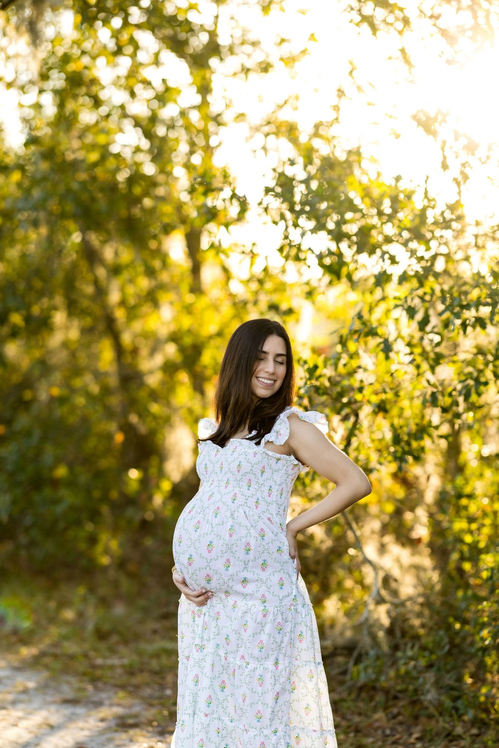 Smiling pregnant woman in floral dress holding baby bump outdoors in golden sunlight surrounded by lush green trees.