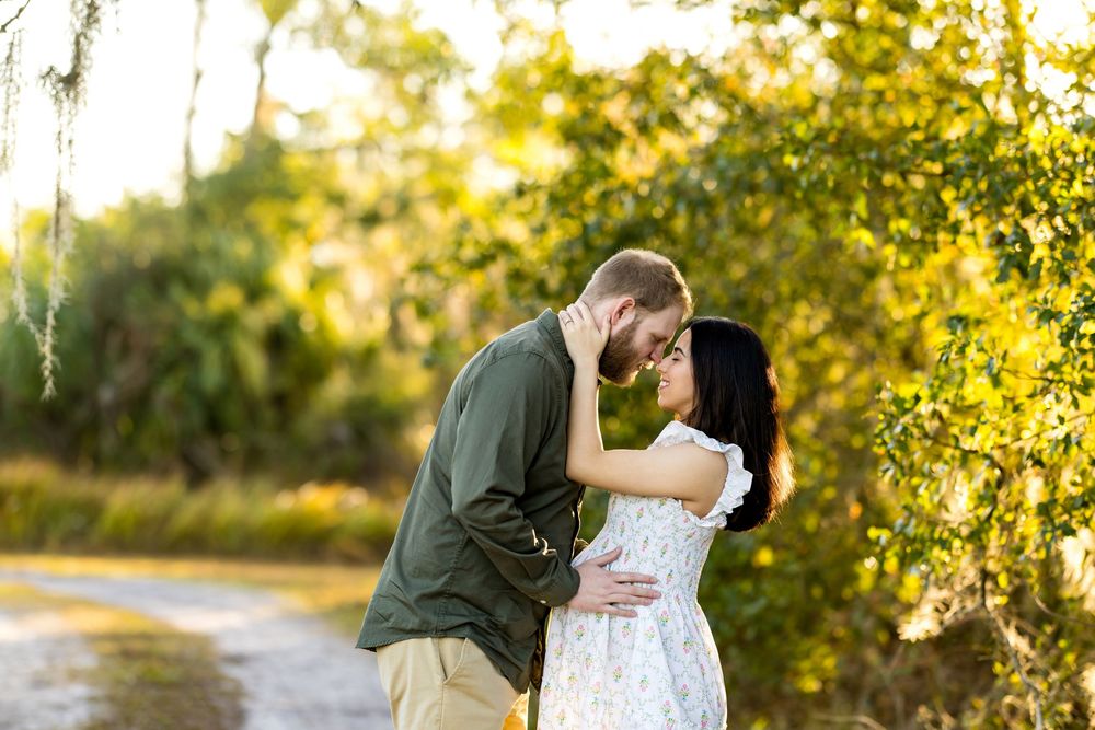 Couple sharing a romantic moment on a sunlit path surrounded by lush green trees during an outdoor engagement session.