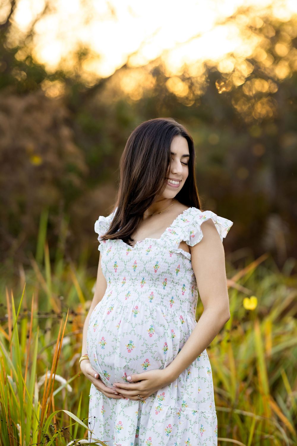 Pregnant woman in white floral dress holding baby bump outdoors at golden hour sunset.