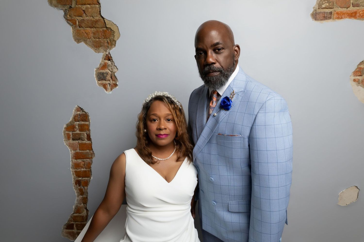 Couple in formal attire poses together, woman in white dress and man in light blue suit, against rustic brick wall.