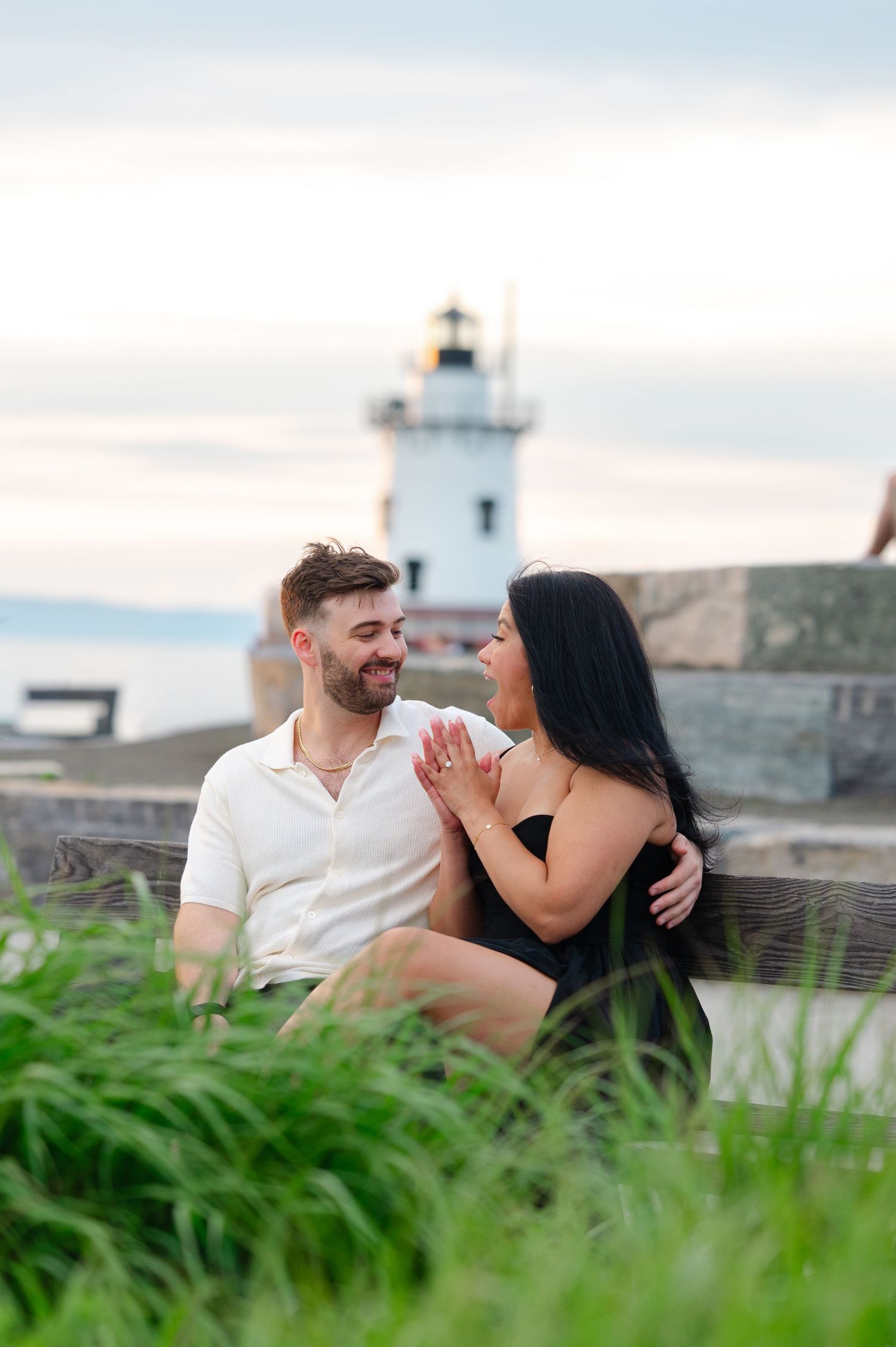 Couple sitting on a bench near a lighthouse, smiling at each other, with lush green grass in the foreground.
